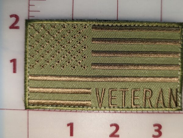 Green embroidered patch with the American flag and the word "VETERAN" in bold text, shown against a gridded background for scale.
